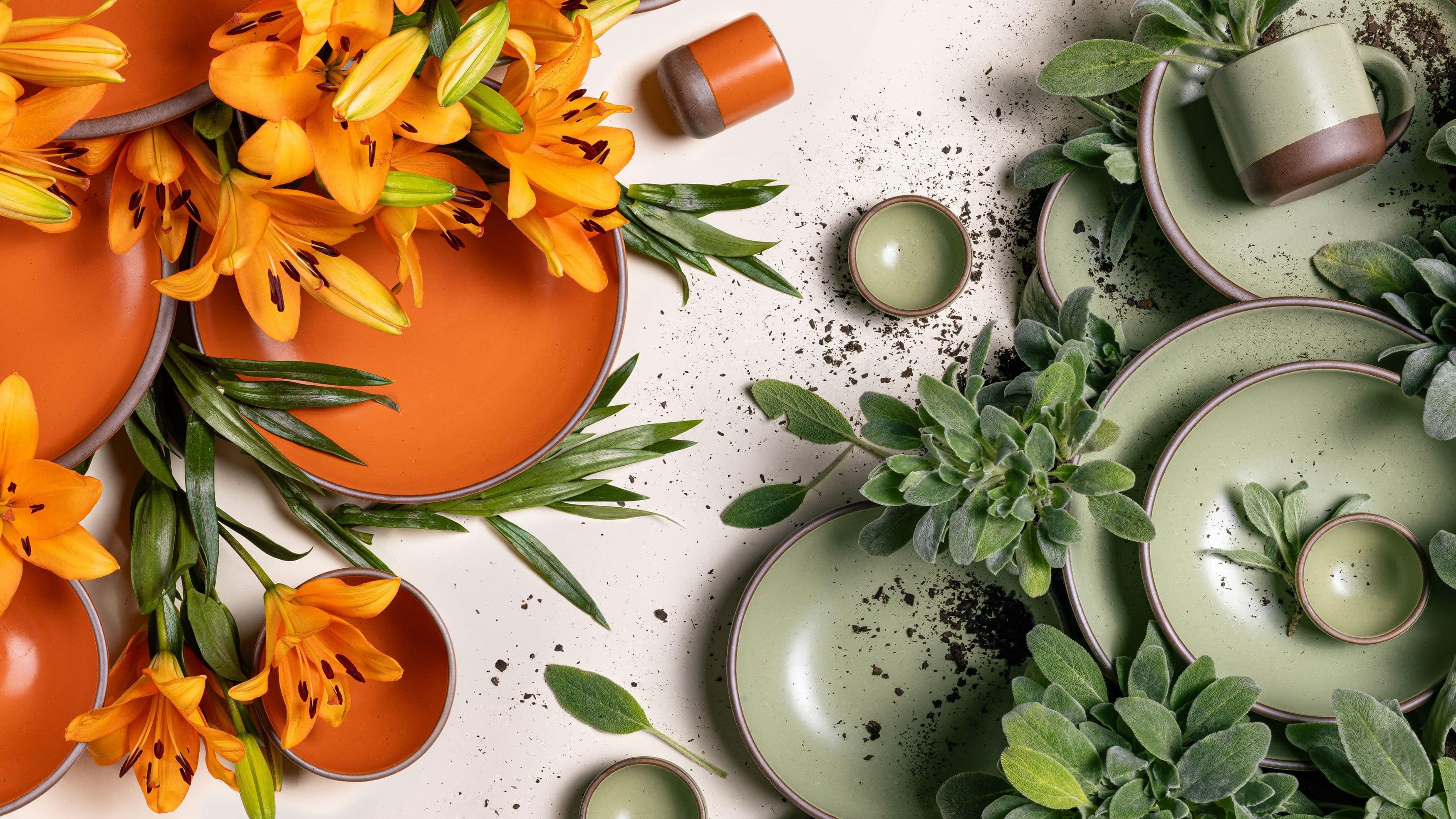 An artful overhead view of ceramic plates, bowls, and cups in a bold orange and calming sage green surrounded by daylilies and lamb's ear. The bold orange pottery and daylilies are on the left, the sage green pottery and lamb's ear on the right.