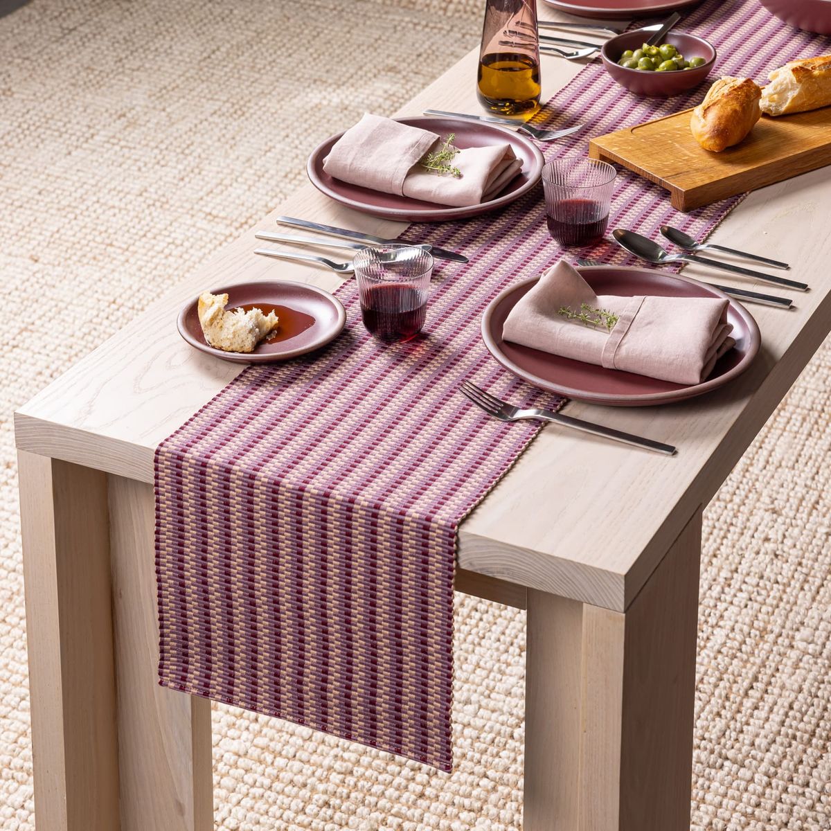 A striped woven runner in plum tones lays on a table with dishes, glasses, napkins, and silverware.