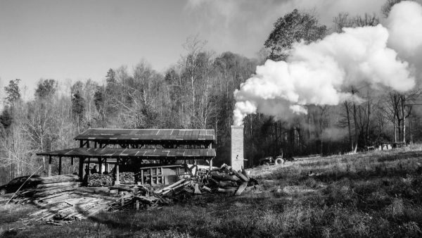 A black and white photo of the wood kiln structure with smoke coming out of the chimney with the woods in the background