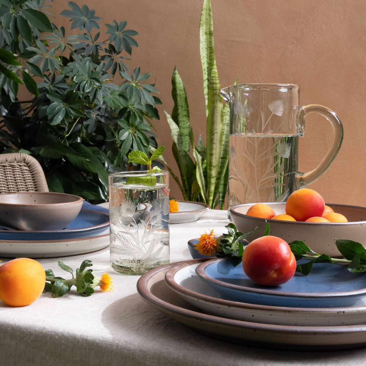An etched thistle tumbler glass and pitcher filled with water, on a table with ceramic dinnerware and fruit, with plants in the background.
