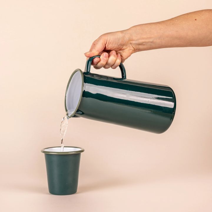 A hand holding a shiny dark green cylindrical jug, with white interior, a spout on the left, and a handle on the right. The jug is pouring water into a dark enamel cup.