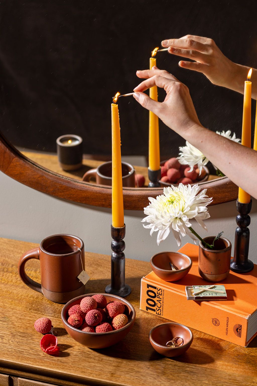 On a table, a hand lights a taper candle sitting in an iron holder. Also on the table are a few small ceramic bowls, and a mug in a cool terracotta color. 