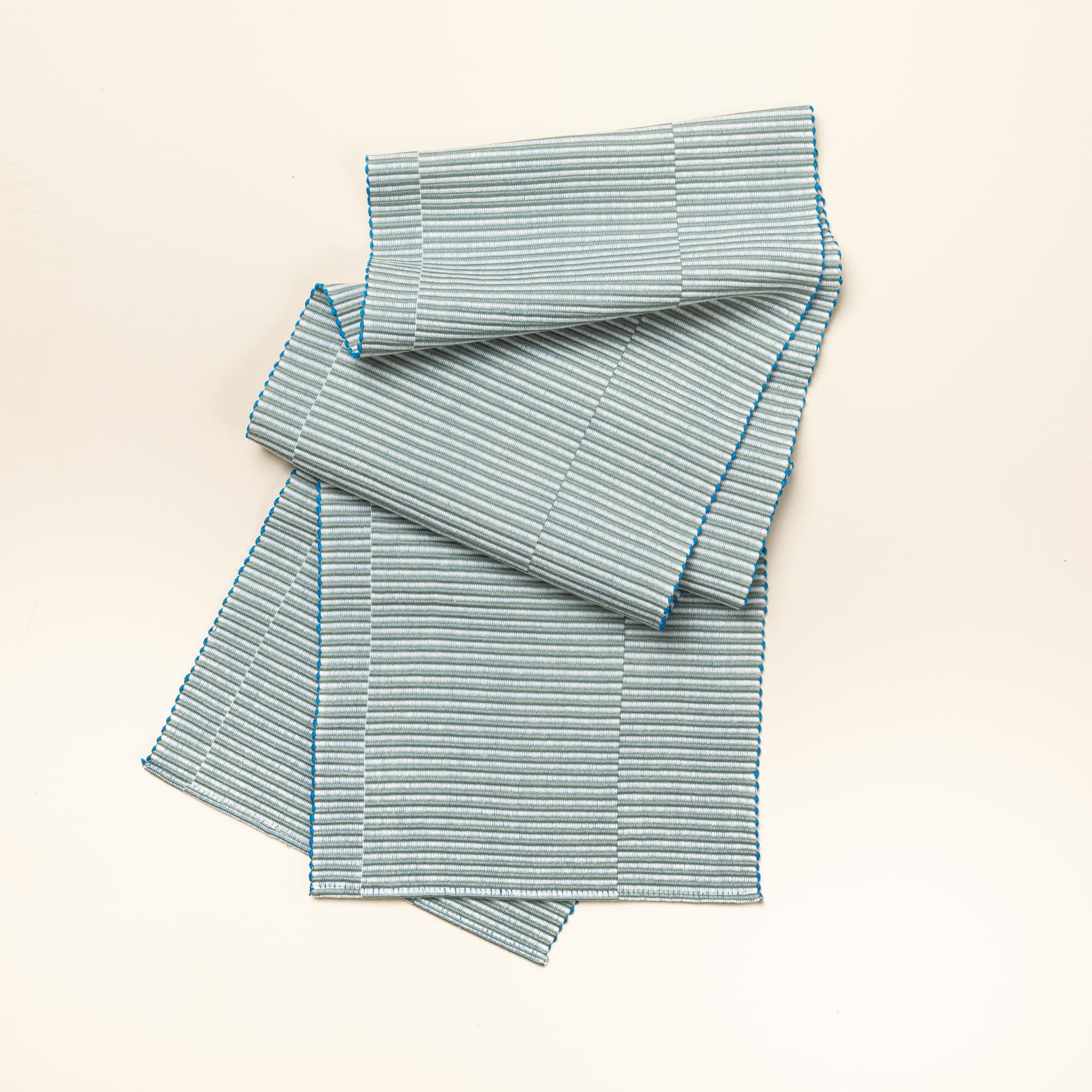 A table runner with offset stripe design that is hand woven in sage and blue colors.
