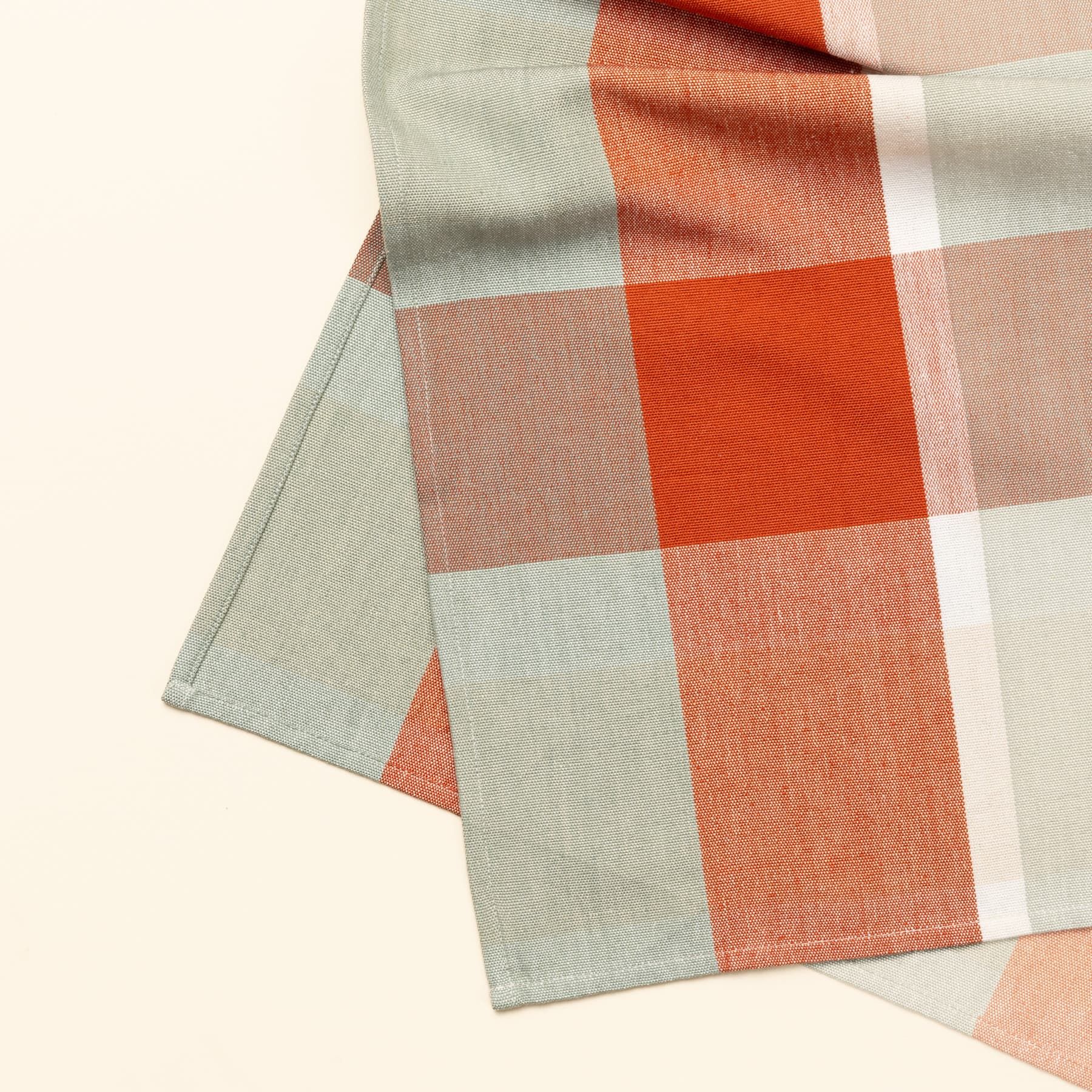 A close up of a corner of a gingham table runner featuring colors of sage green, bold orange, and cream.