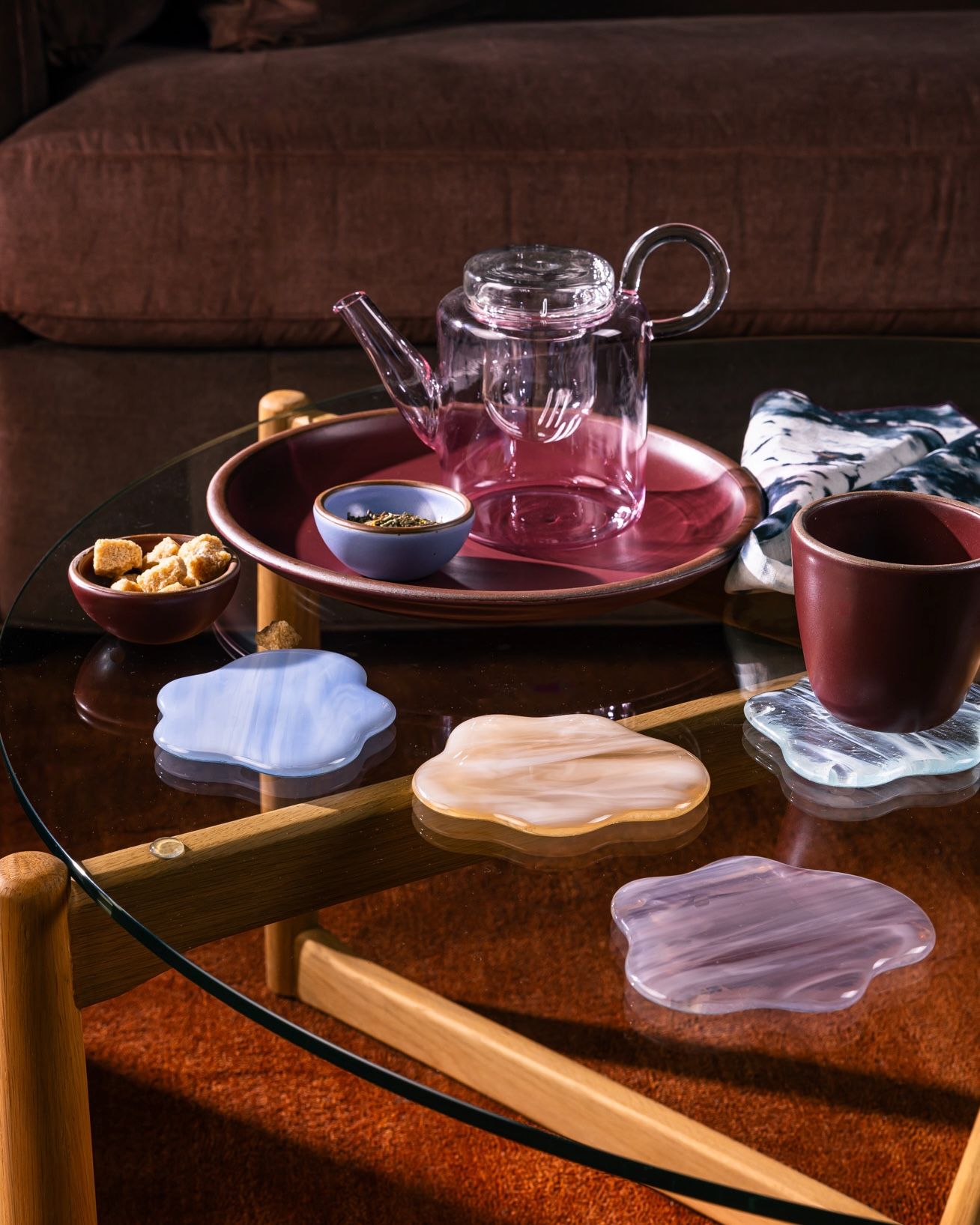On a coffee table, there is a pink glass teapot, coasters shaped like clouds, and ceramic cup and plate in a warm plum color