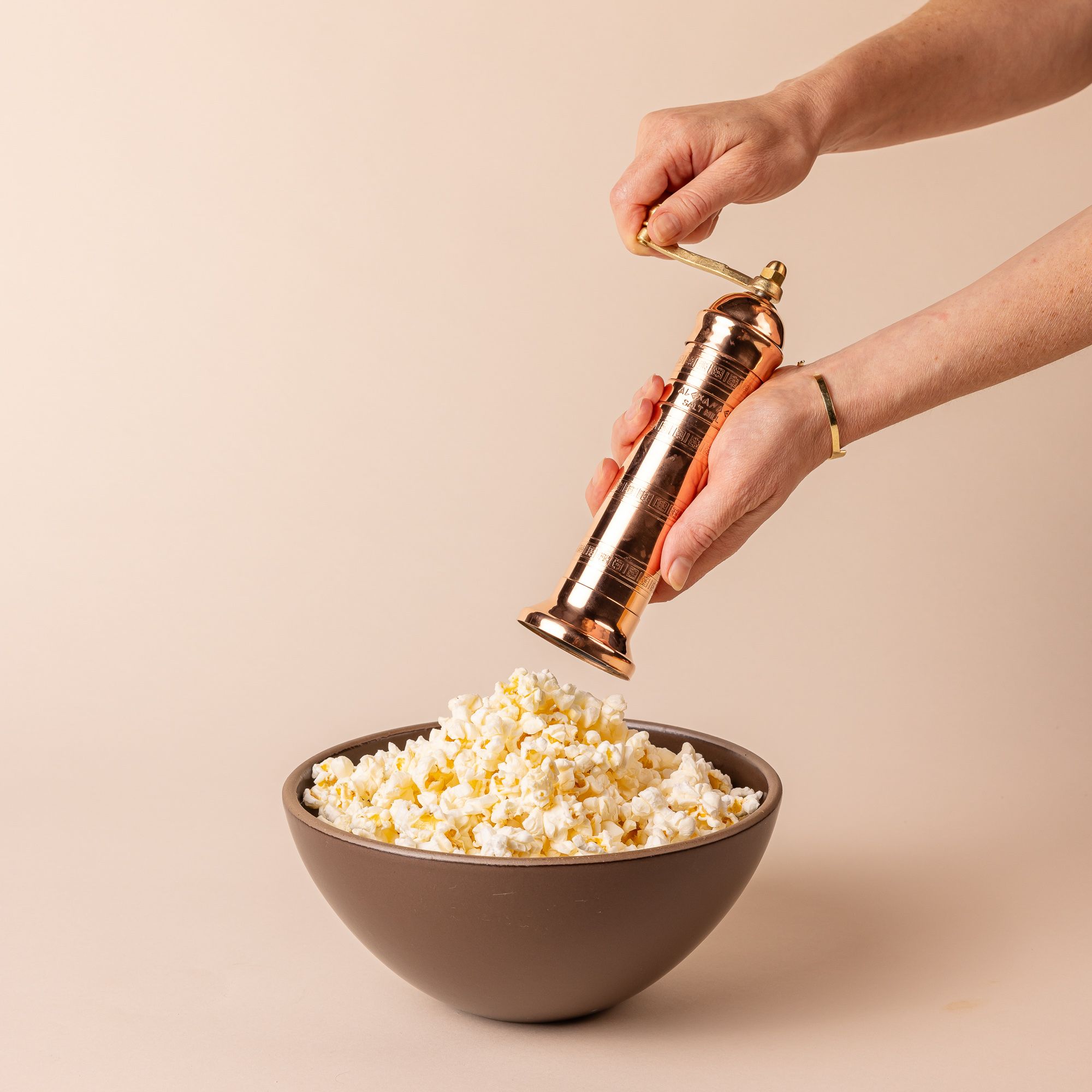 A hand holding a narrow all copper salt mill with a flanged base and rotating the grinder above a bowl of popcorn.