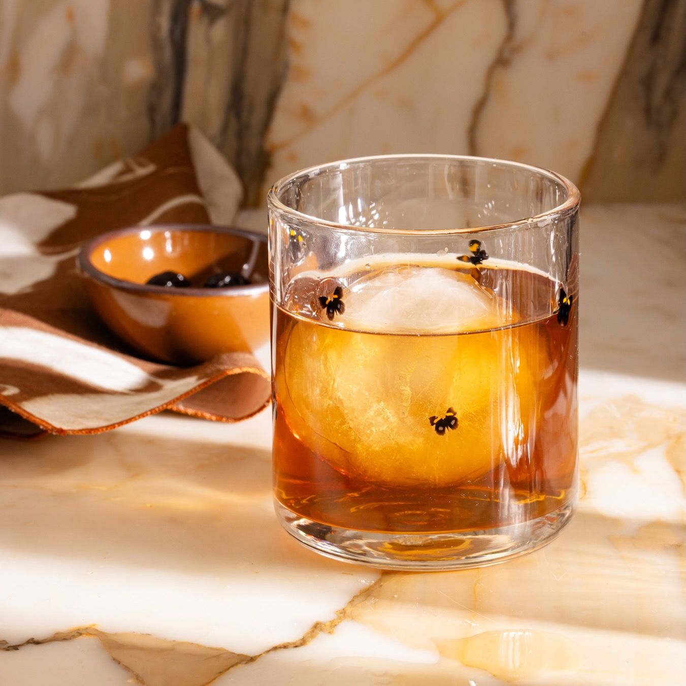 Whiskey cocktail in a clear glass with butterfly motifs, served over a large ice sphere on a marble table.