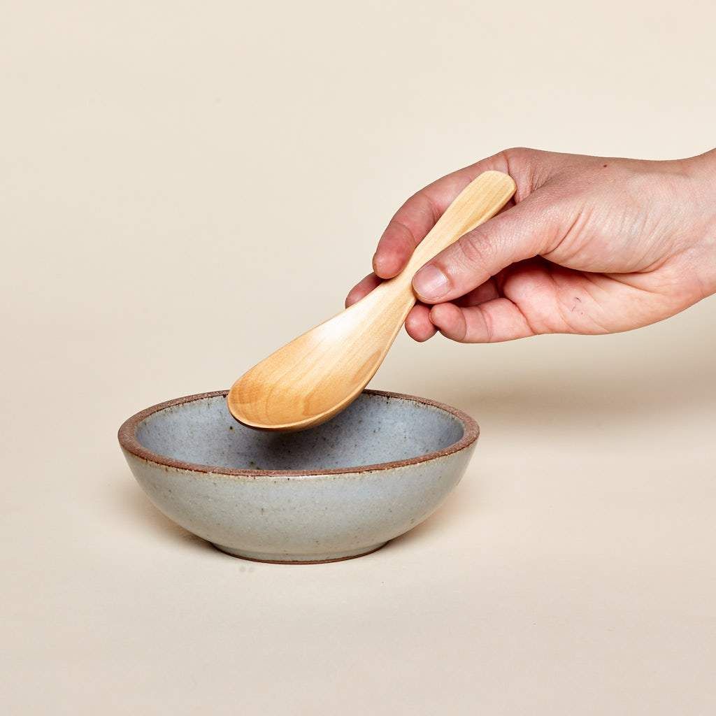 Hand holding a wooden soup spoon that measures 6 inches long above a small bowl