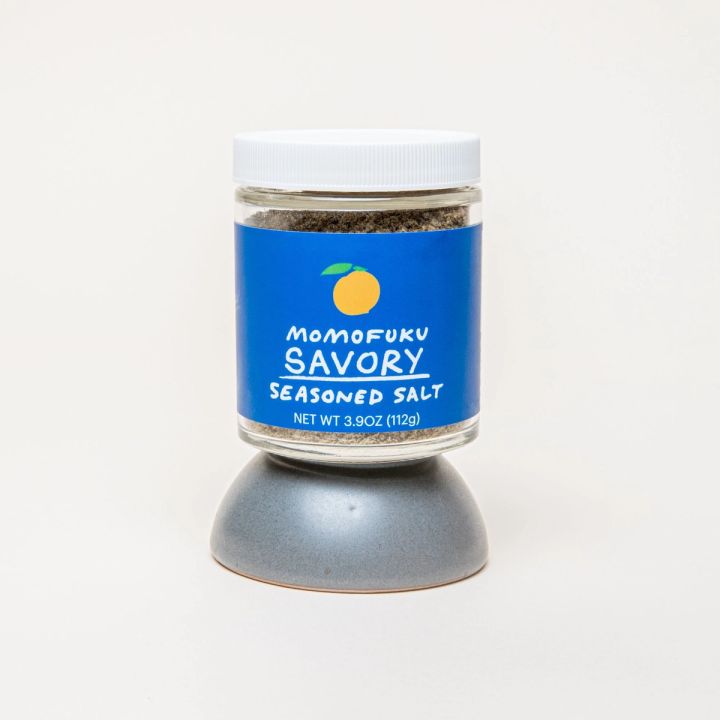 A jar with a white top and blue label that reads "Momofuku Savory Seasoned Salt", sitting on top of an upside down tiny grey bowl