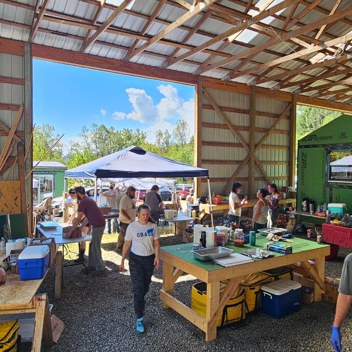 People are gathered doing different tasks in a barn-like space filled with tables, a pop-up tent, and supplies