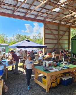 People are gathered doing different tasks in a barn-like space filled with tables, a pop-up tent, and supplies