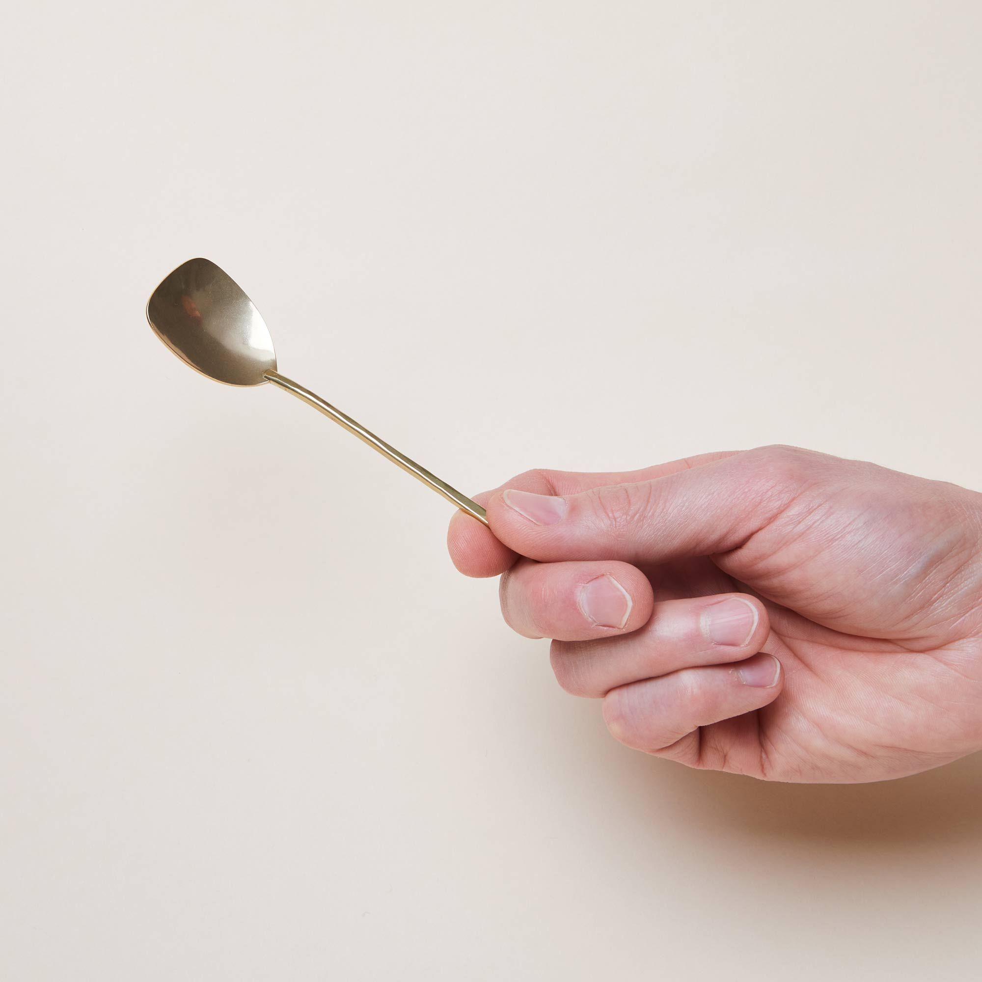 A hand holds a brass spoon with a flared and squared-off bowl attached to a long, thin brass handle