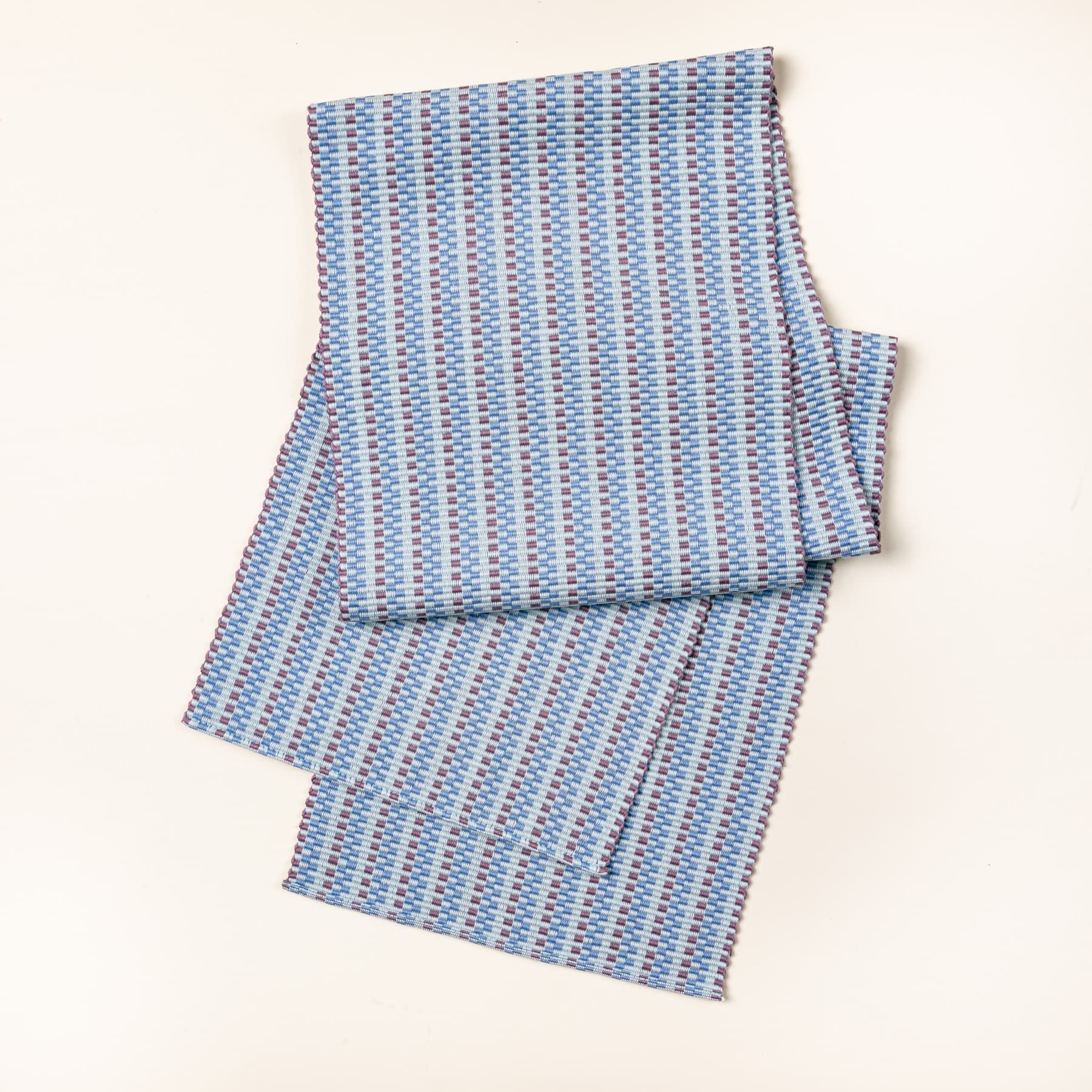 A folded woven table runner with thin horizontal striped soft blues and purple colors