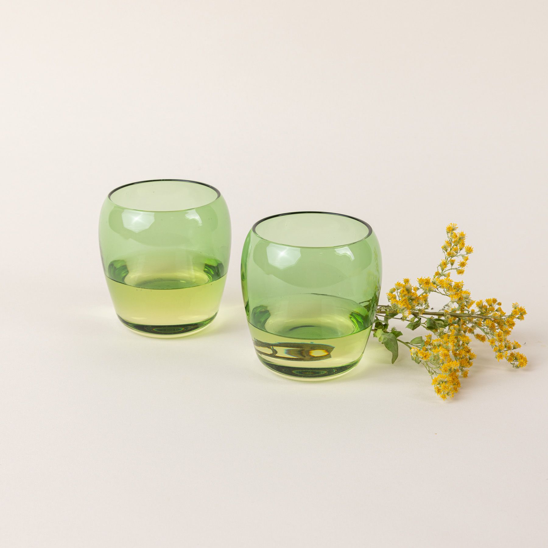 Two short light greenglass tumblers with a slight curve inward on the top. The glasses are filled with white wine and a flower is sitting behind them.