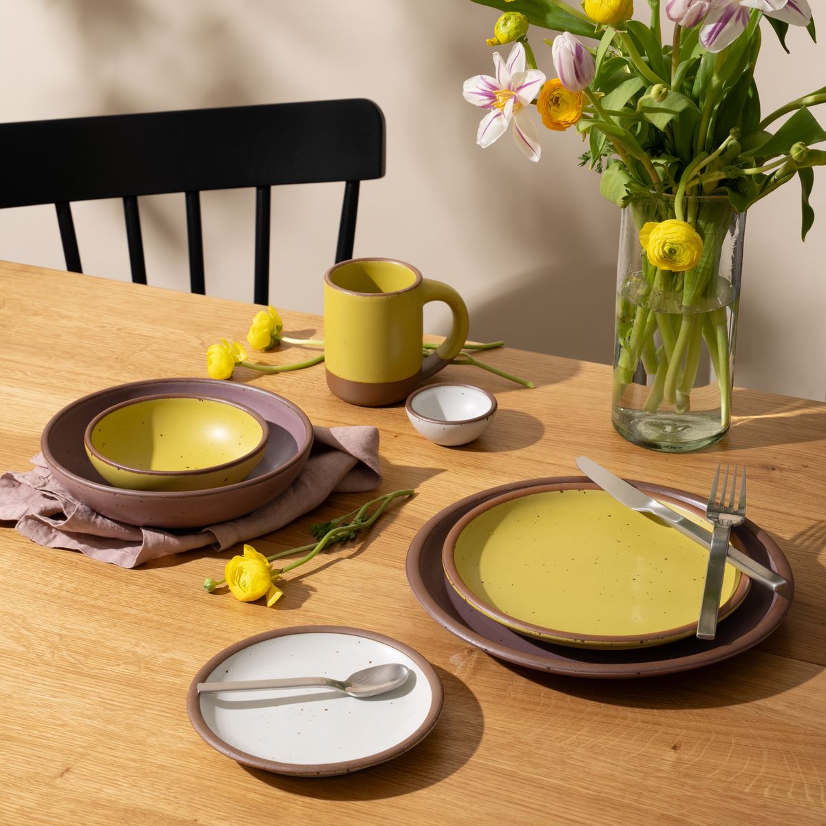An artful dinnerware set featuring a mug, a bitty bowl, 3 different sized plates and 2 small and large bowls in soft mauve purple, zesty yellow and cool white colors on a table. There is also silverware on the plates, a tall glass of water, and a tall clear vase with flowers.