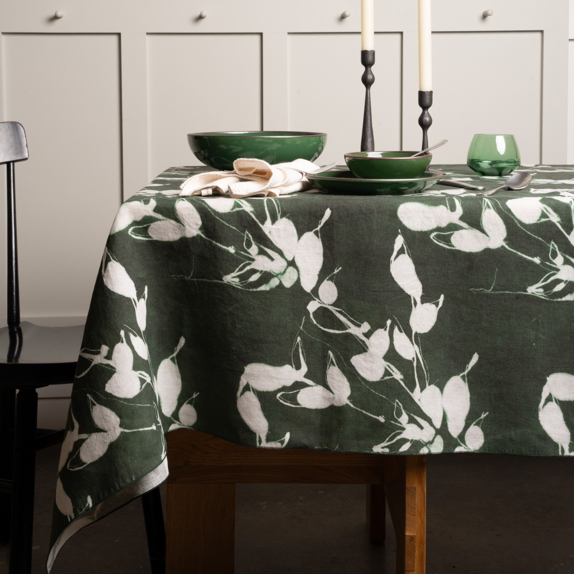 Dark green tablecloth with white botanical print, set with green dishes, candles, and glassware on a wooden table.