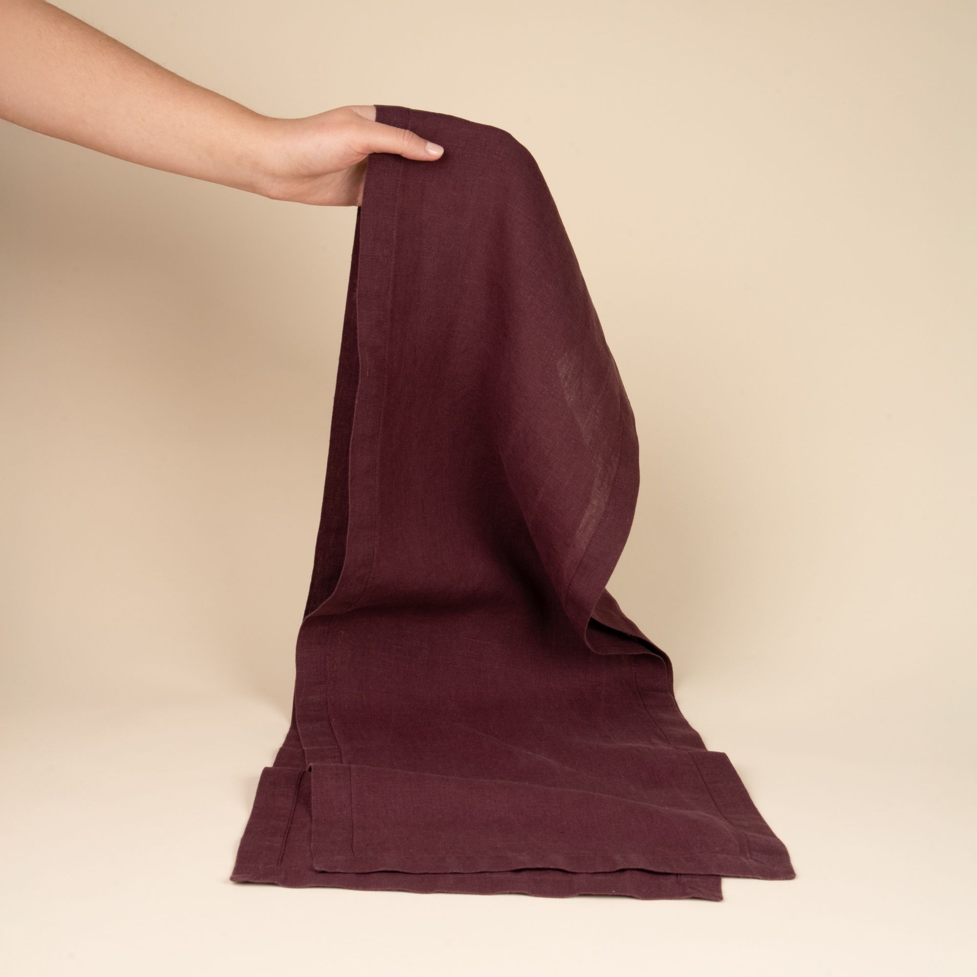 A hand holds up a folded linen table runner in a deep purple color