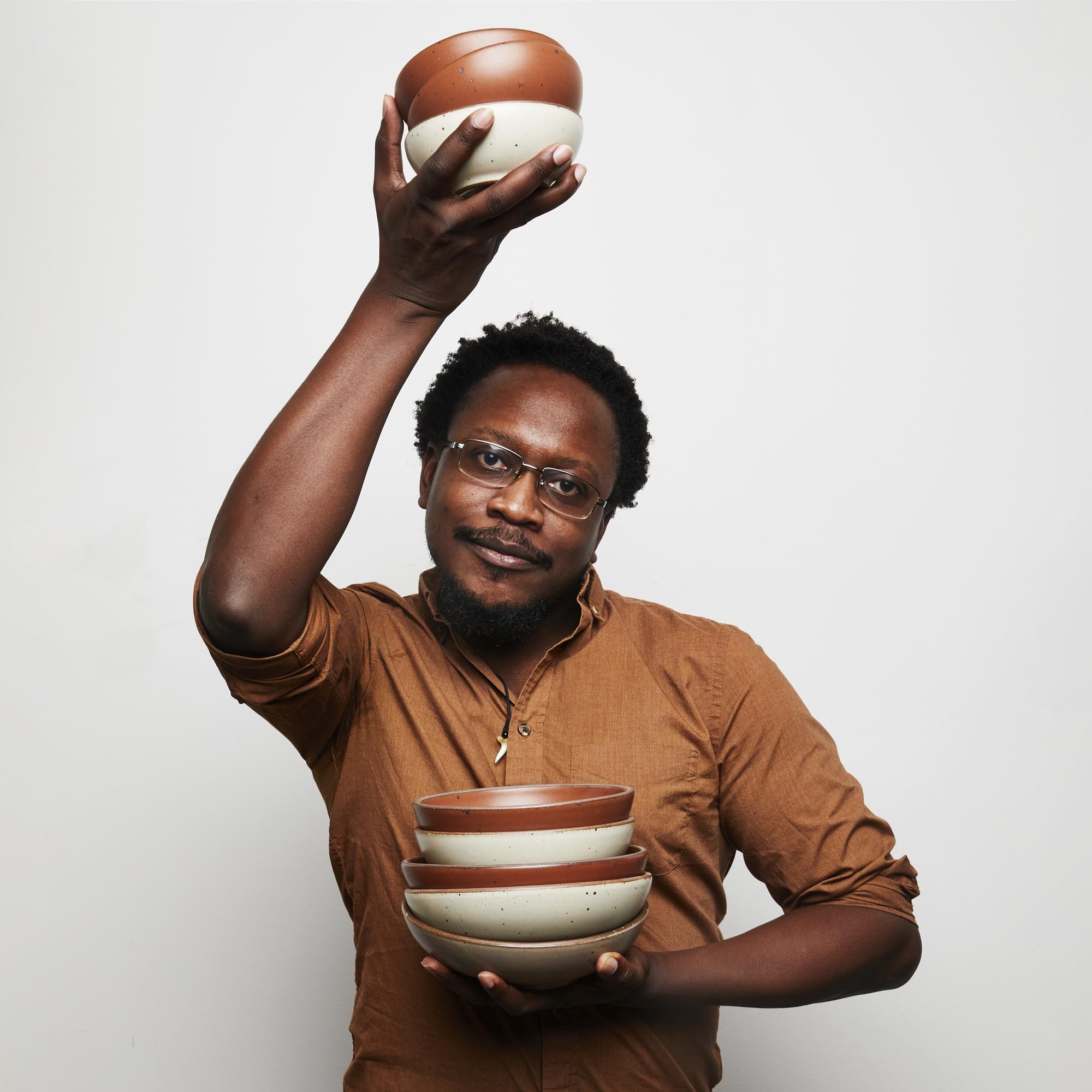 A person wearing glasses holds a stack of bowls close to the chest in one hand and another stack of bowls above his head in the other hand.