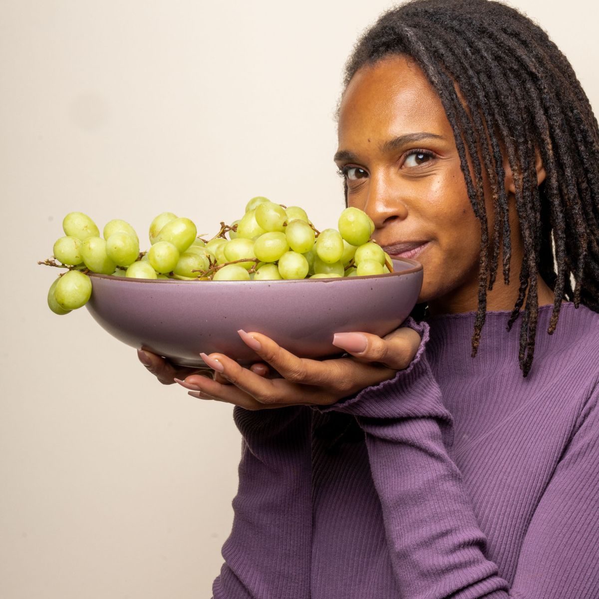 A woman is slightly smiling and staring at the camera holding a large shallow serving bowl in soft mauve purple, holding green grapes.