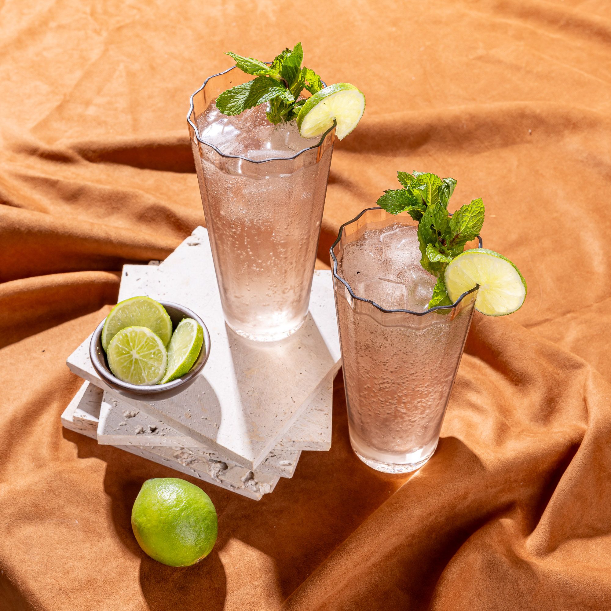 Two tall transparent light amber glasses with wide grooves on the side. The glasses are filled with a cocktail with lime and mint and styled with nearby lime and lime slices..