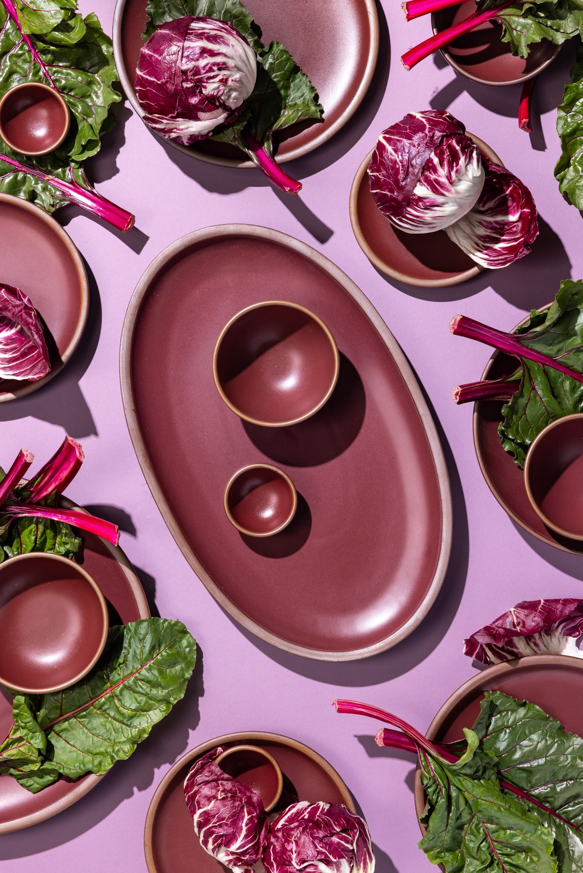 Ceramic dinnerware in a deep plum color against a light colored table, with stalks of colorful leafy green and purple veggies