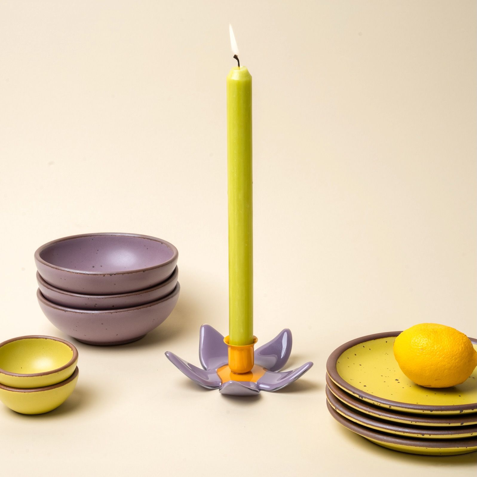 A lit taper candle in a neon green color in a orange and purple flower candleholder, surrounded by zesty yellow and soft mauve purple dinnerware.