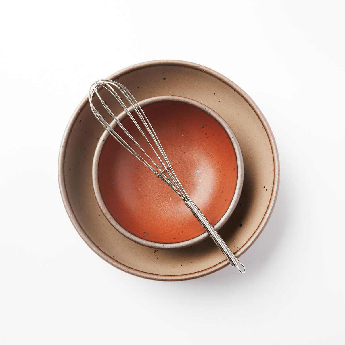 A tiny slim stainless steel whisk lays on top of 2 stacked bowls in terracotta and warm light brown