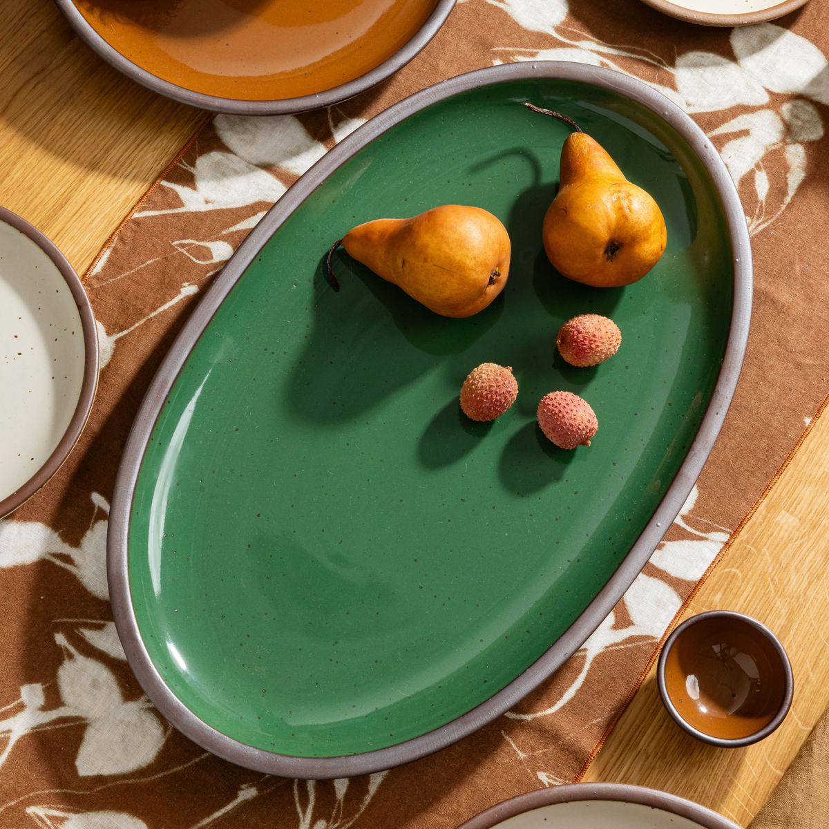 Glossy emerald green oval platter with two pears and three lychees on a patterned table runner with matching dishes.