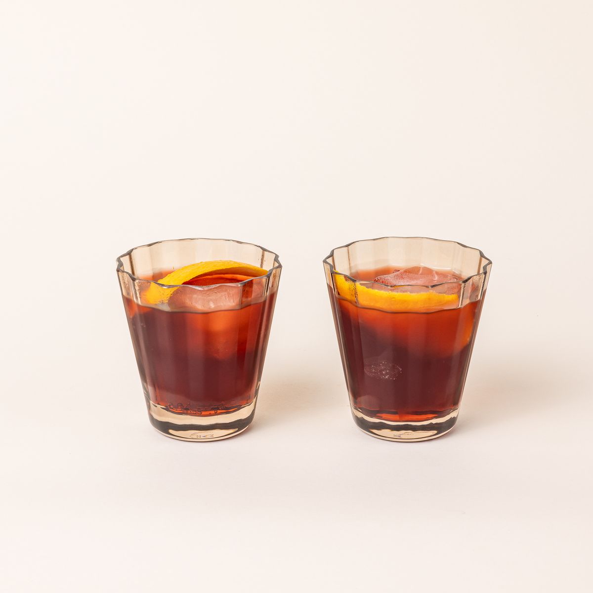 Two transparent light amber glasses with wide grooves on the side, filled with a cocktail and orange slice.