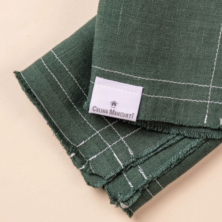 A folded deep teal pair of linen napkins with fringe edges and a white maker's tag.
