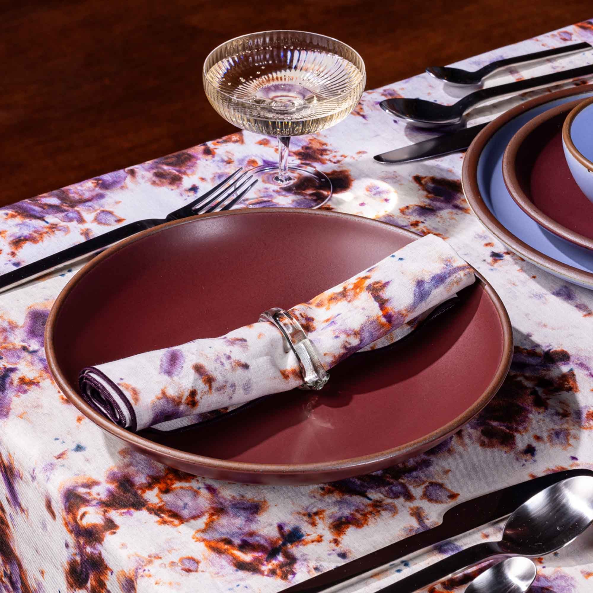 A rolled linen napkin in a marbled pattern with white, pink, purple, and orange colors sits on a ceramic plum dish.