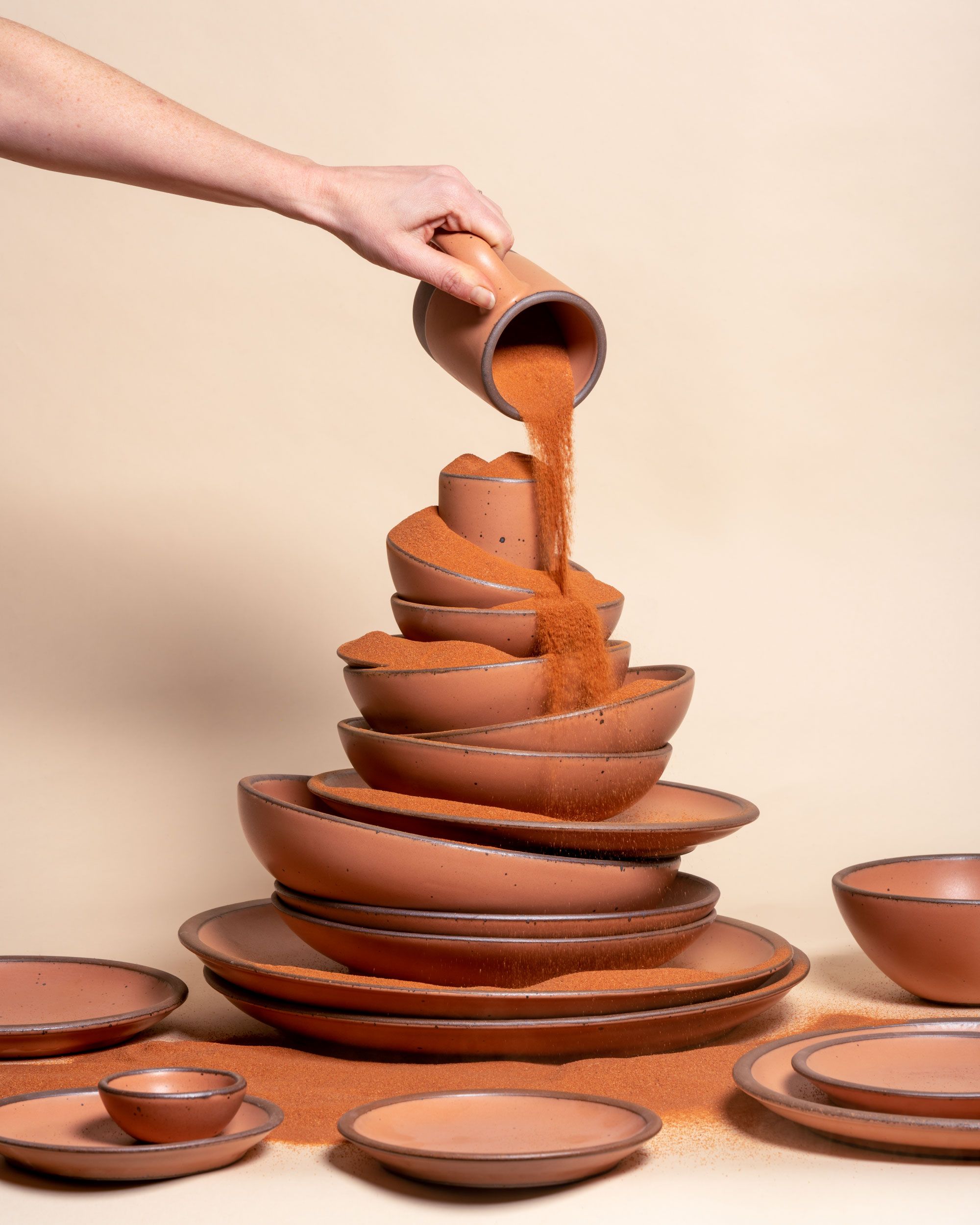 A stack of nude plates and bowls. Overhead, a hand holds a mug and pours a similarly colored desert sand out of a mug on top of the stack of pottery.