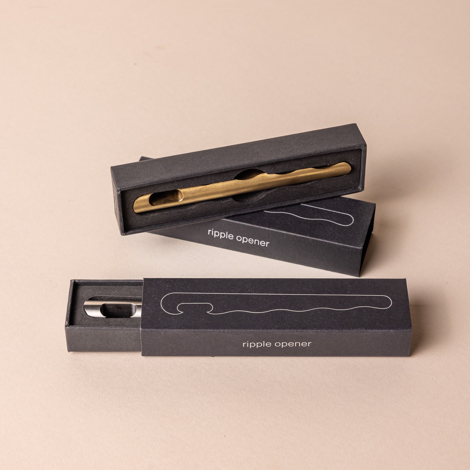 Two black boxes that hold sophisticated brass and steel bottle openers with a simple design and a wave with grooves all along the opener side.