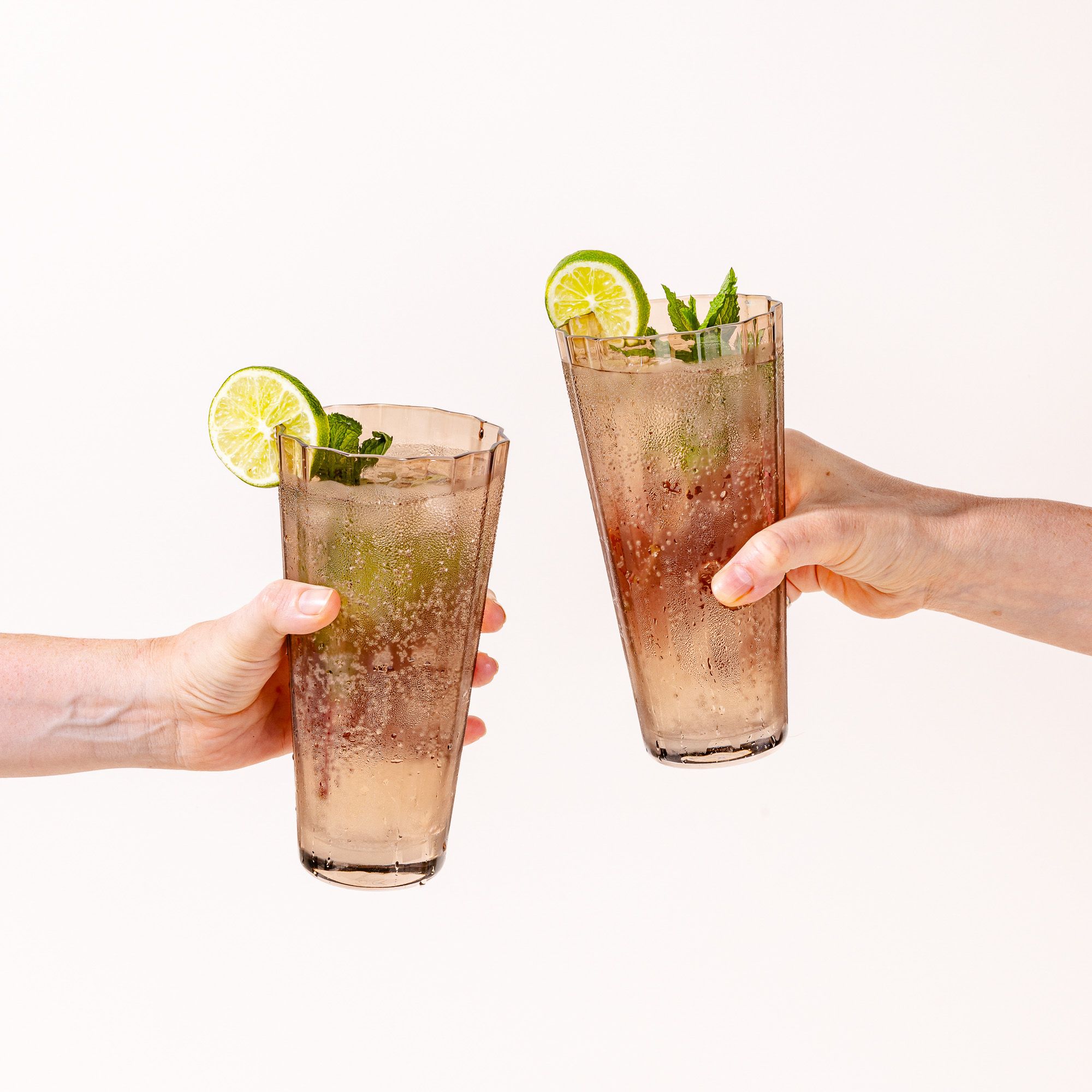 Two hands are cheering two tall transparent light amber glasses with wide grooves on the side. The glasses are filled with a cocktail with lime and mint.