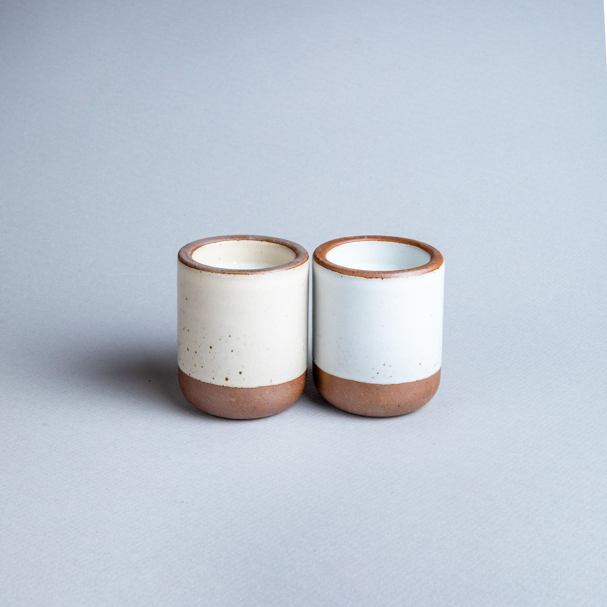 Two small ceramic vessels in cream off-white and cool white colors with candles inside each.