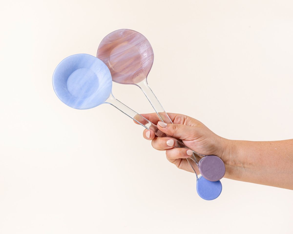 A hand holds  modern glass colorblocked serving spoons in periwinkle and plum colors.