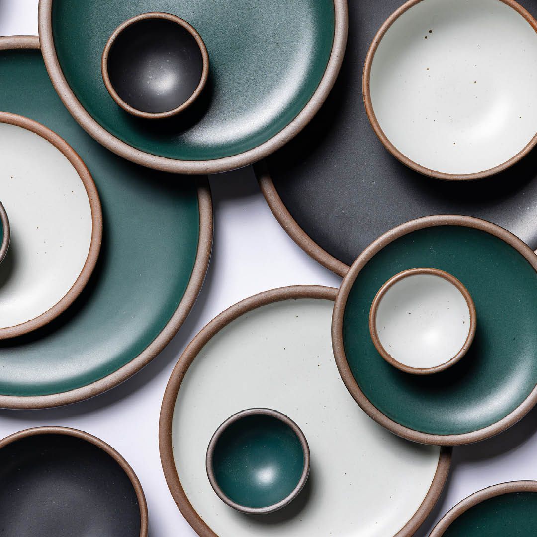 Overhead view of ceramic plates and bowls of various sizes and in dark teal, white and graphite colors stacked on each other.