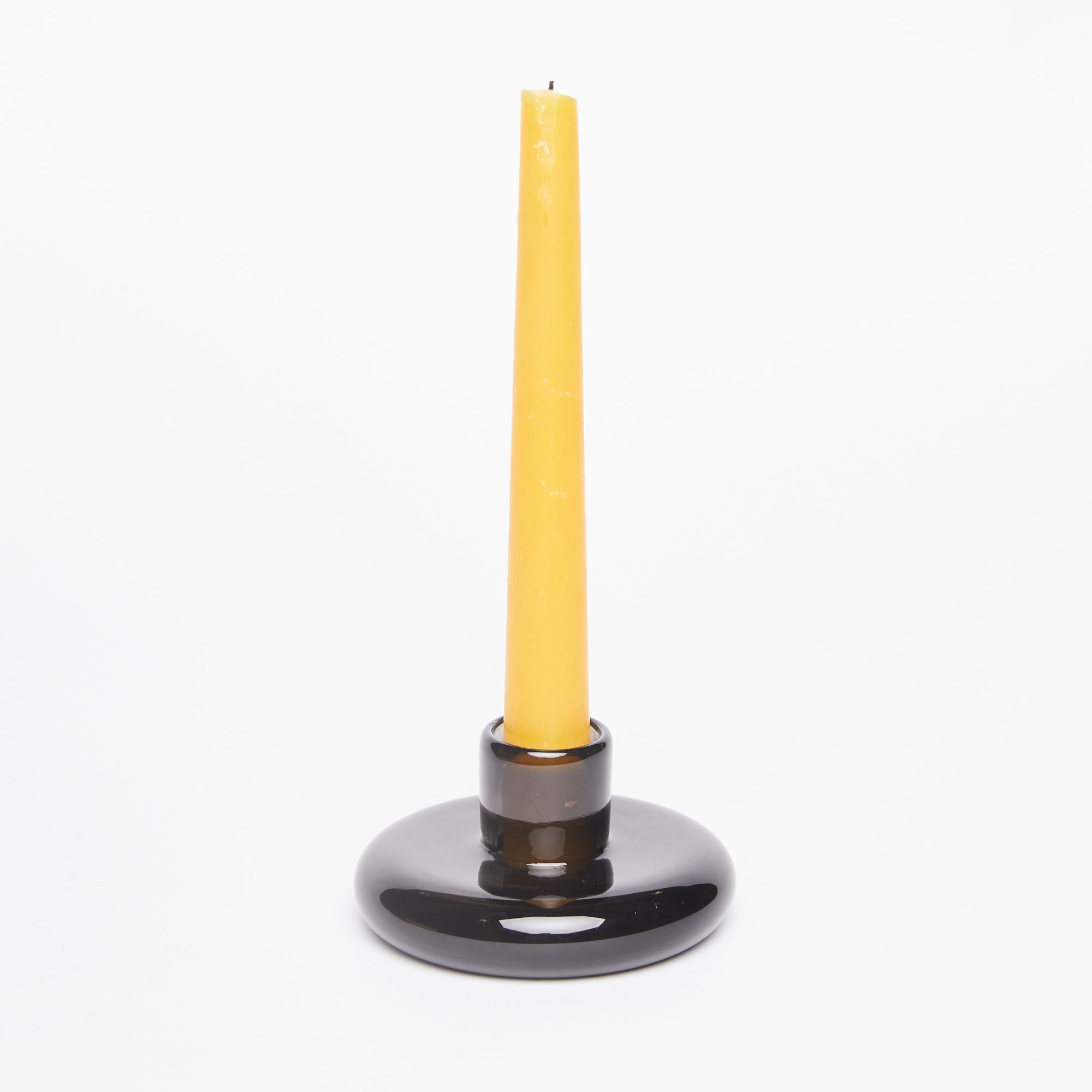 Yellow taper candle sitting in a candle holder made of a clear dark grey glass disc-like base with a small cup for the candle at center