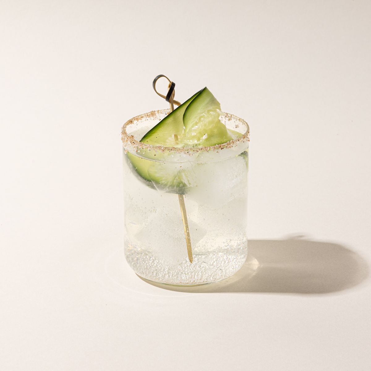 A straight walled short glass cup with a clear cocktail and cucumber garnish.