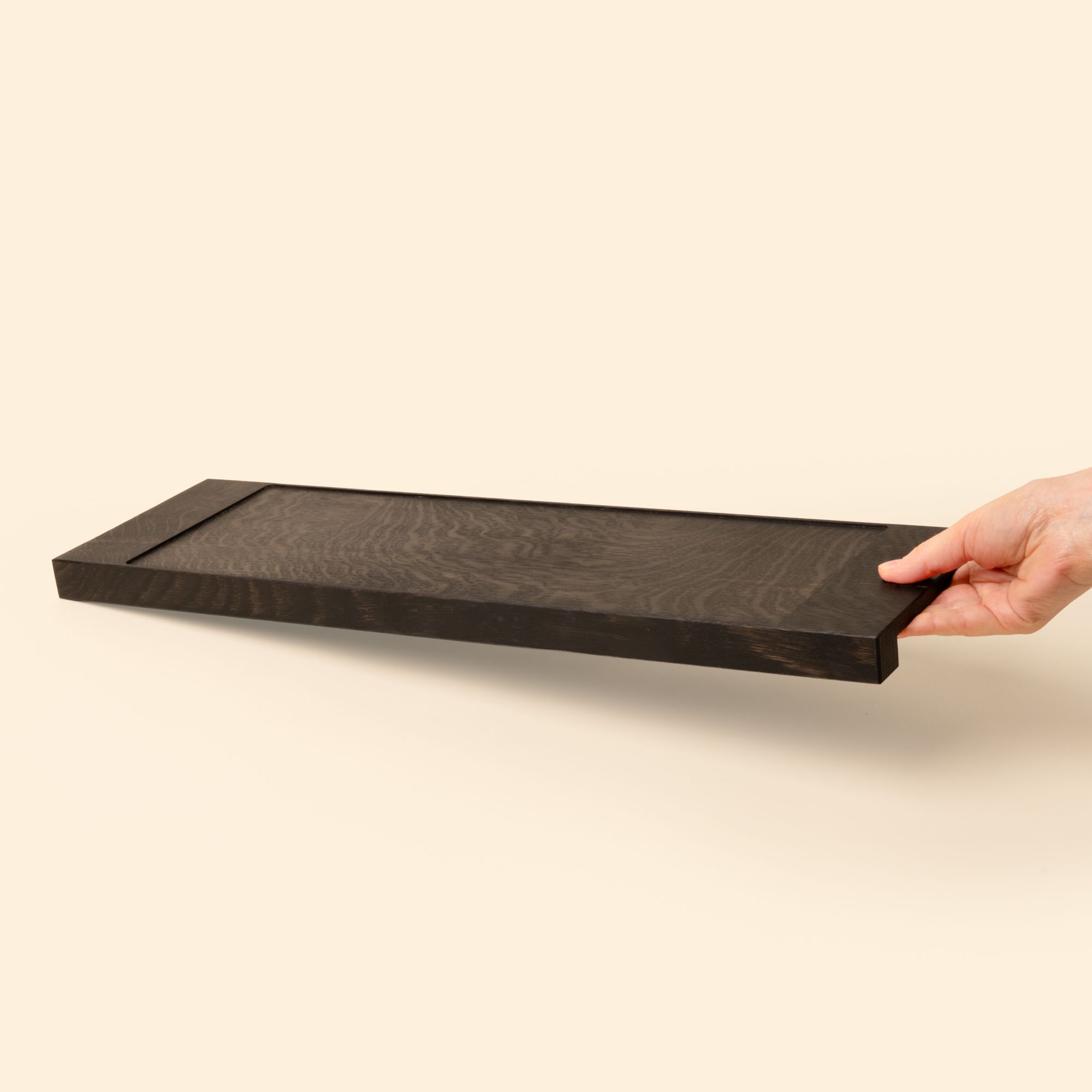 A hand holds out a narrow rectangle of ebonized oak wood with a vertical groove at either end, by the beveled handle on the side.