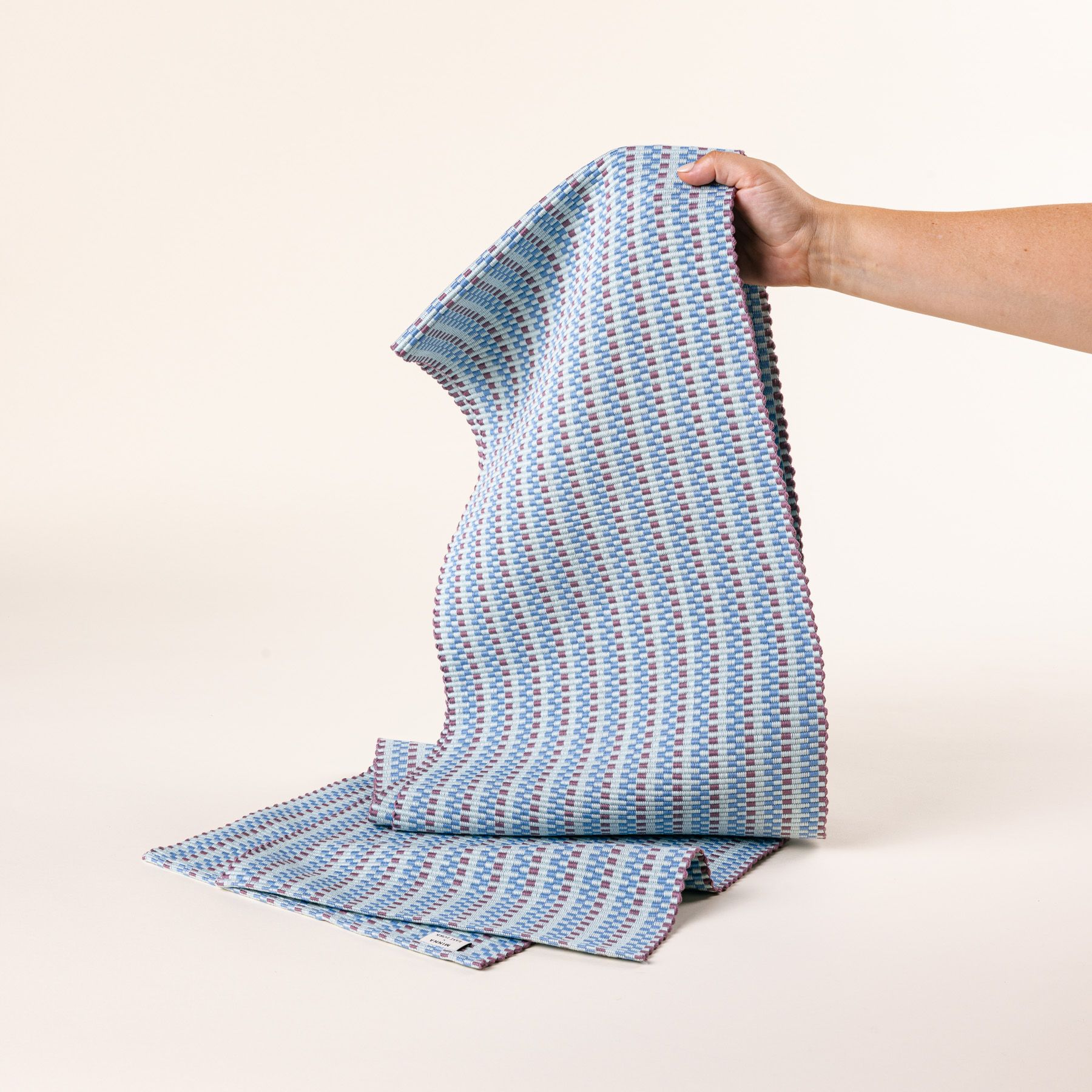 A hand holds up a woven table runner with thin horizontal striped soft blues and purple colors