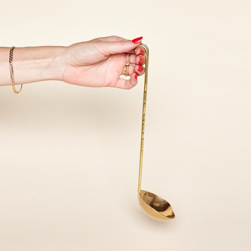 A hand holds the handle of a brass ladle at an angle so some of the bowl's underside is visible.