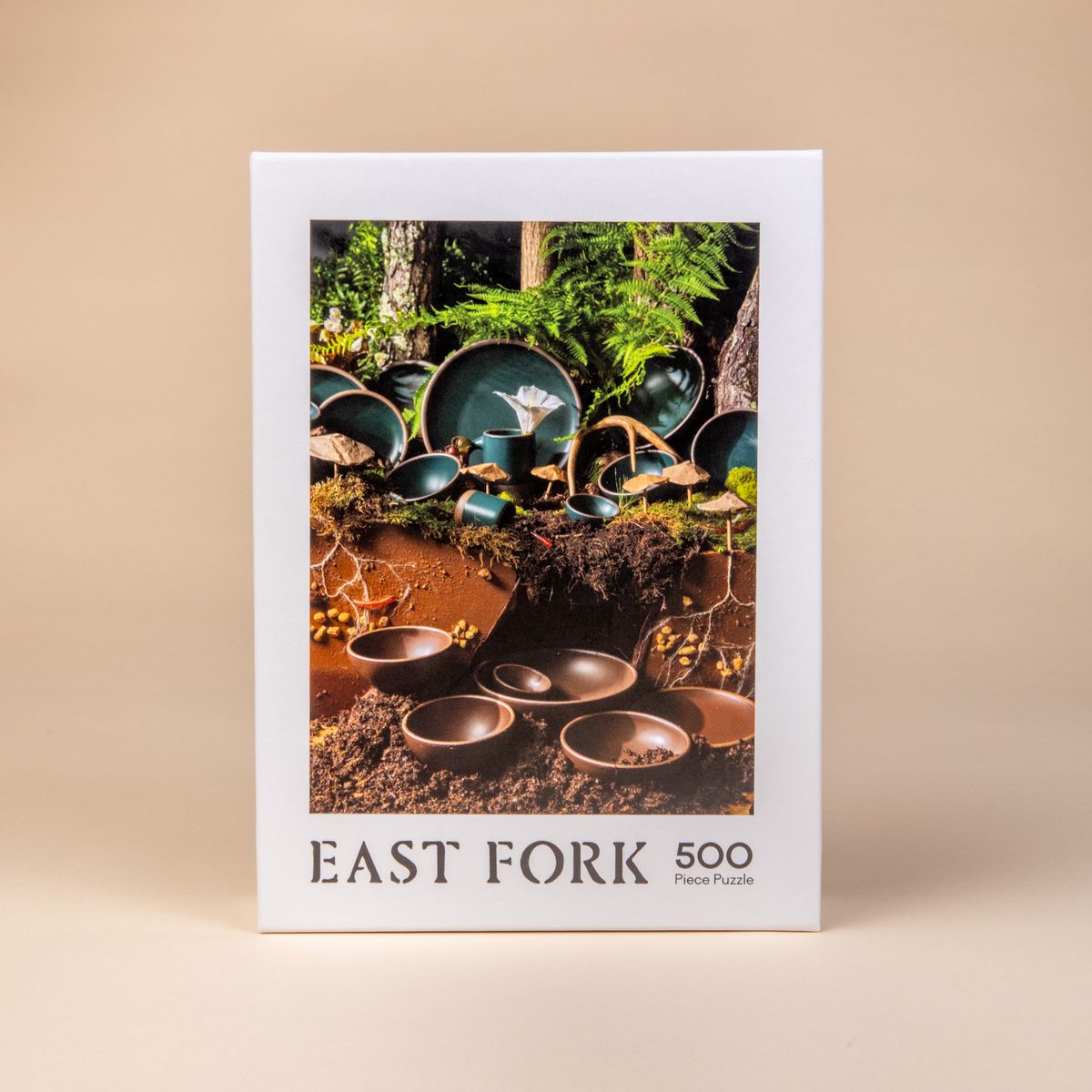The front of the box of a 500-piece jigsaw puzzle that when put together shows ceramic bowls, plates and mugs in dark teal and brown colors in a natural forest setting.