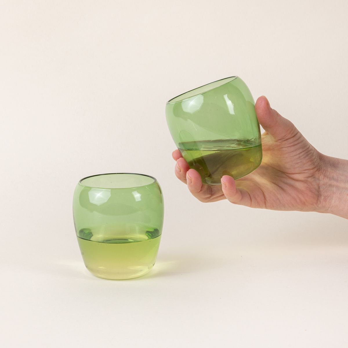 Two short light green glass tumblers with a slight curve inward on the top. A hand is holding the glass on the right.