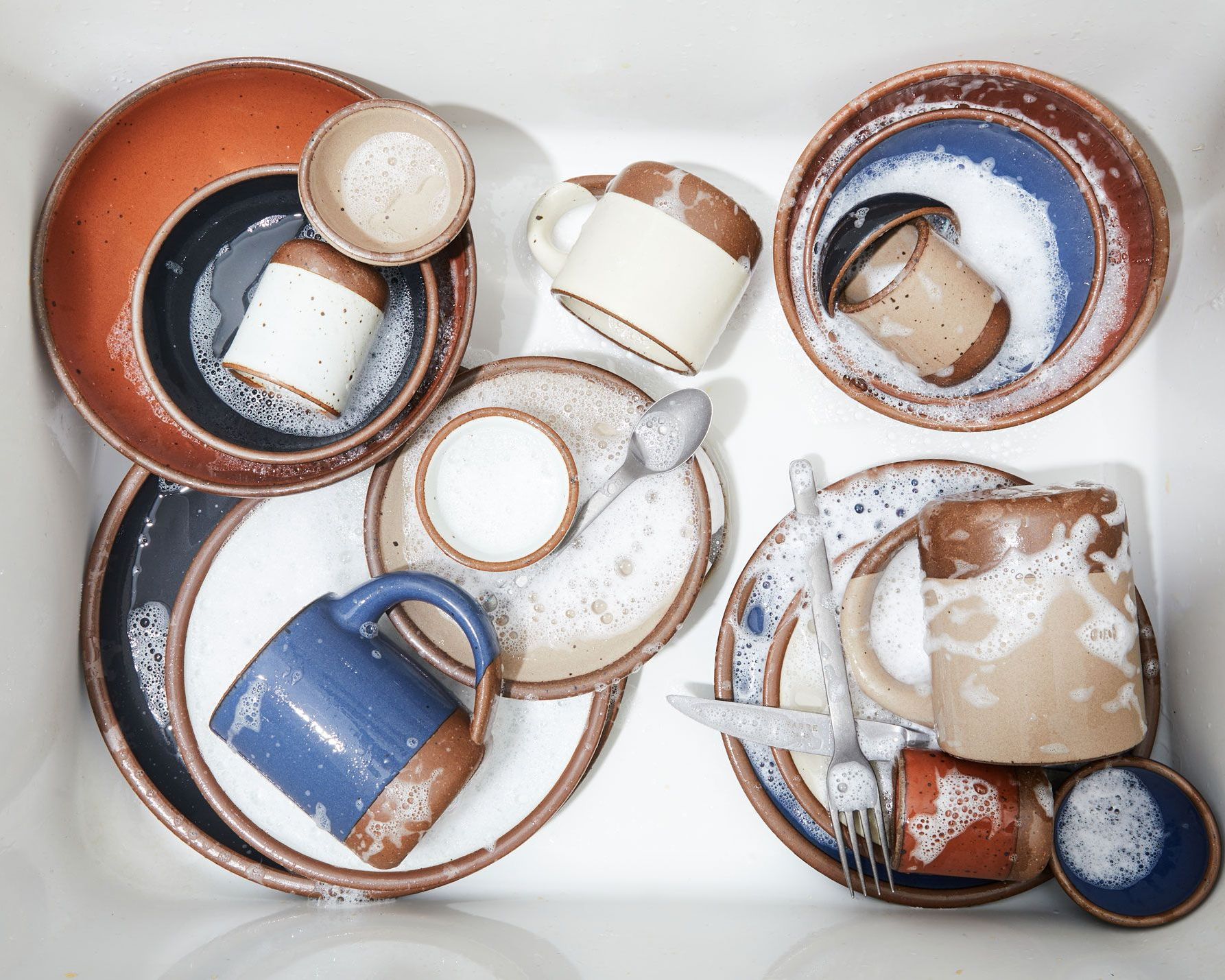 A stack of plates and mugs in core colors (blue, white, black, brown) in a sink covered in soap