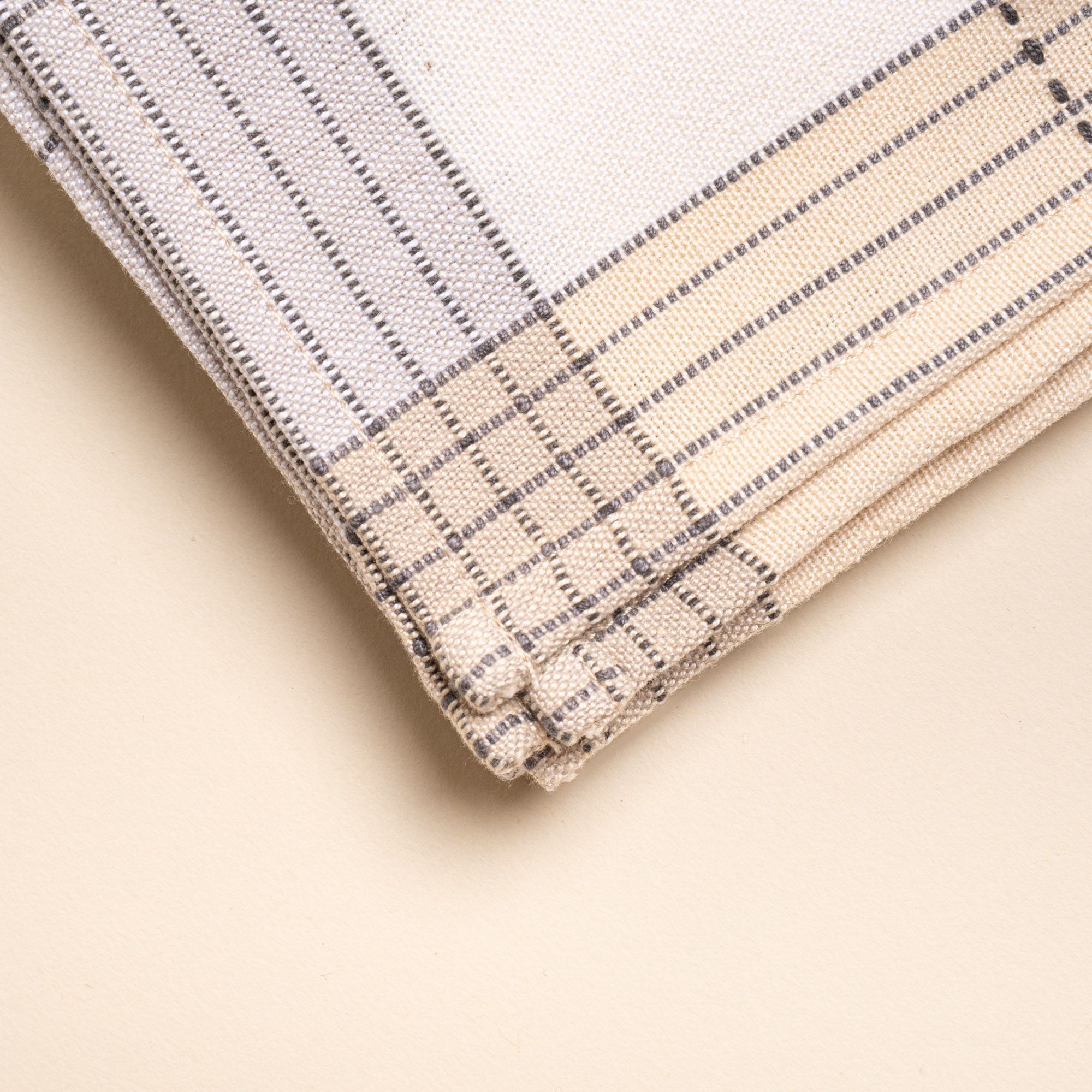 A closeup of the corner of a white napkin with grey gridlines with light grey and natural striped edging