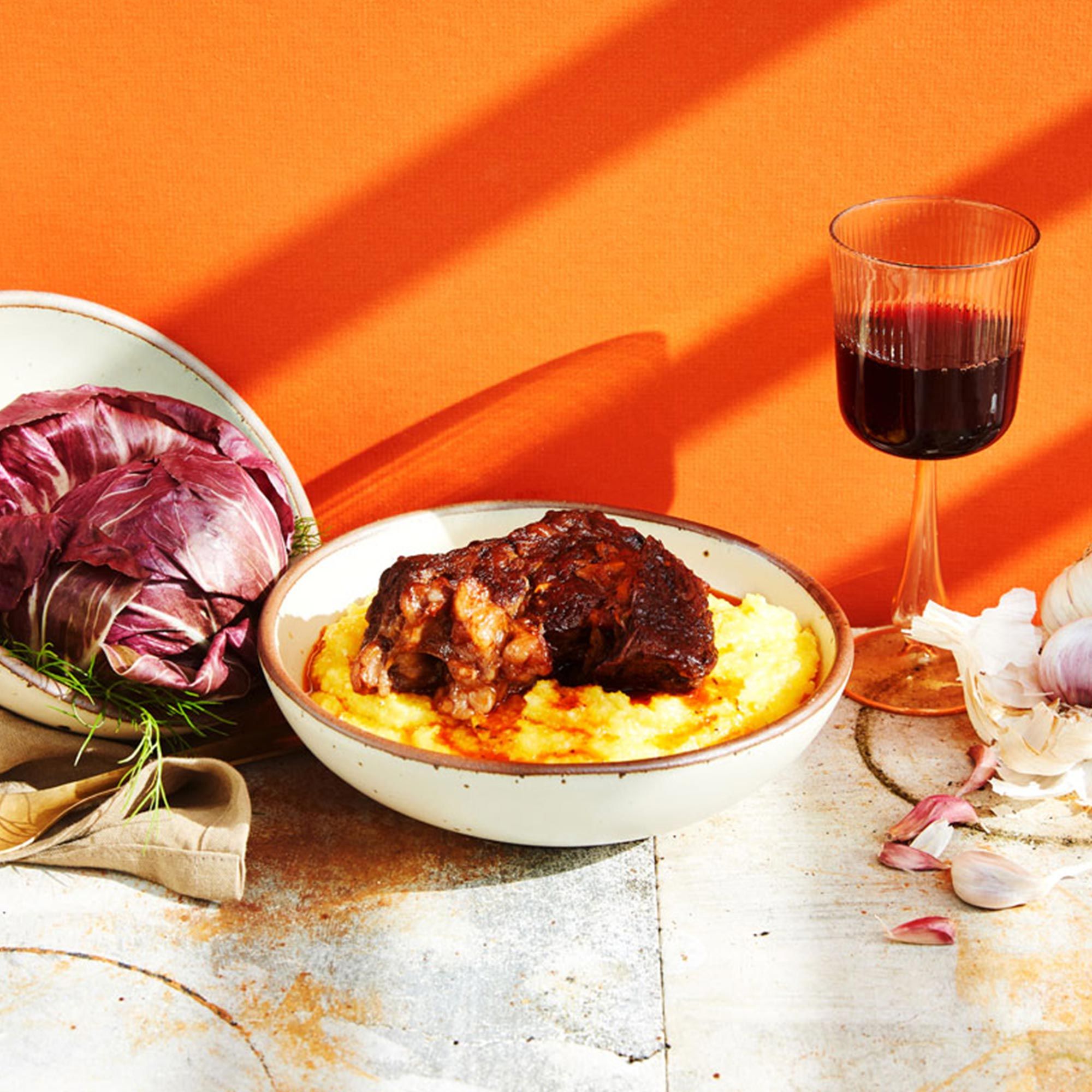A table setting with ceramic dinnerware in a cool white color, plated food, a wine glass with red wine, and onion and garlic surrounding.