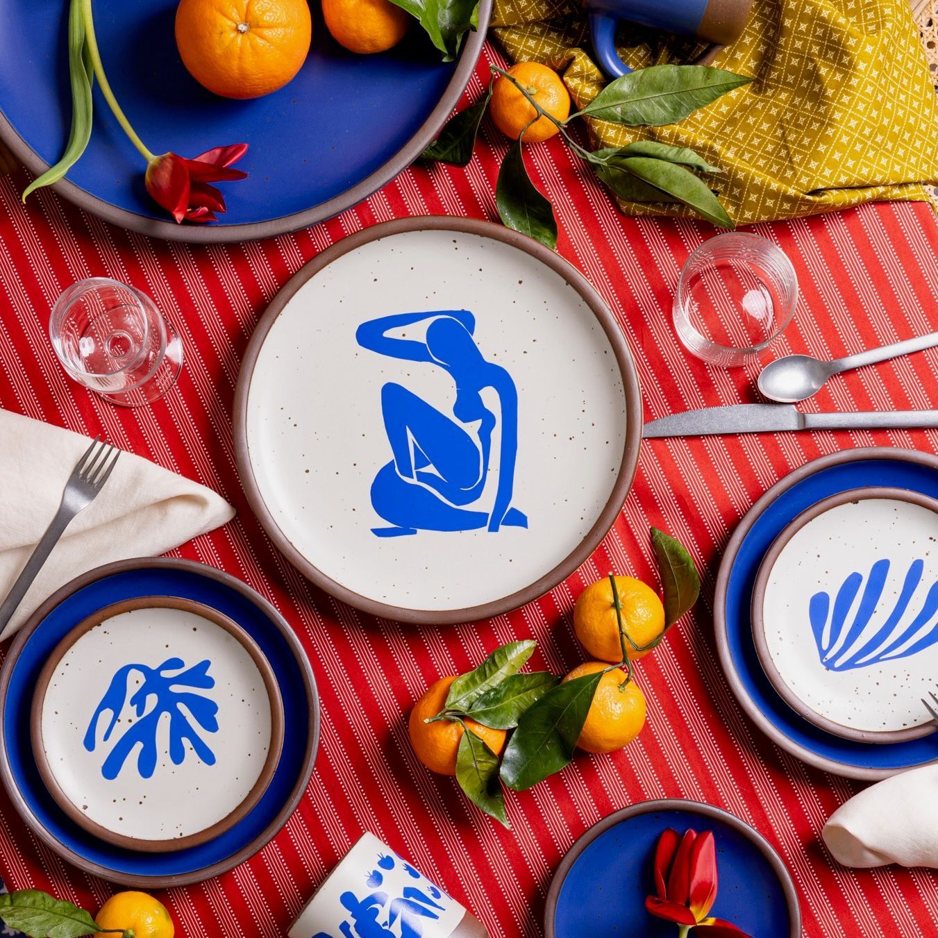 On a colorful table are plates in a bright cobalt blue, with decorated plates featuring artwork by Henri Matisse, along with silverware, glasses, and oranges.