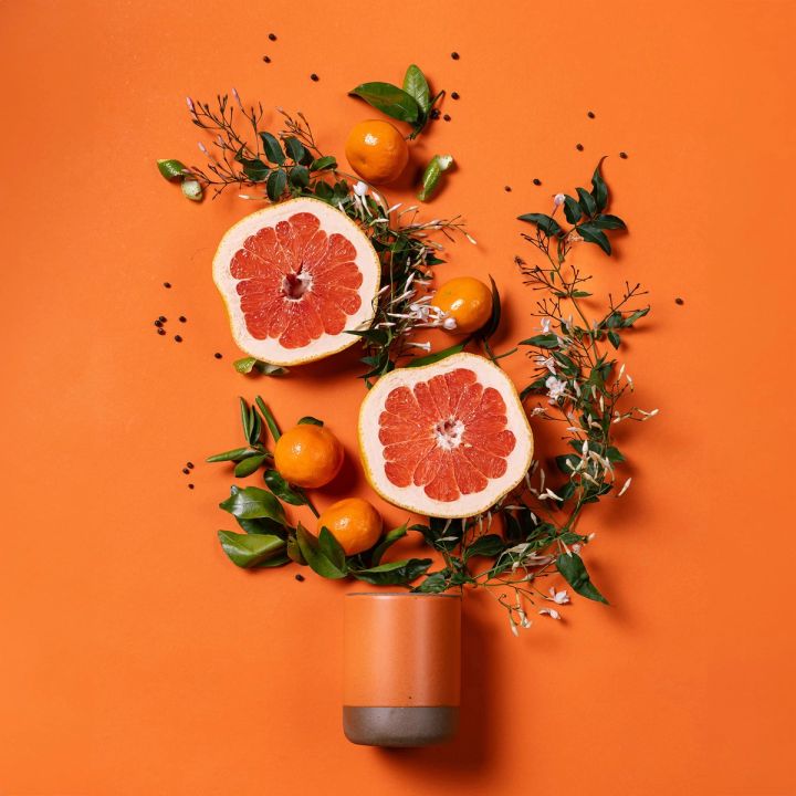 A cylindrical ceramic vessel in a bold orange color laying on its side - sliced grapefruits, oranges, and greenery are artfully styled coming out of the top of the candle to reflect the scent.