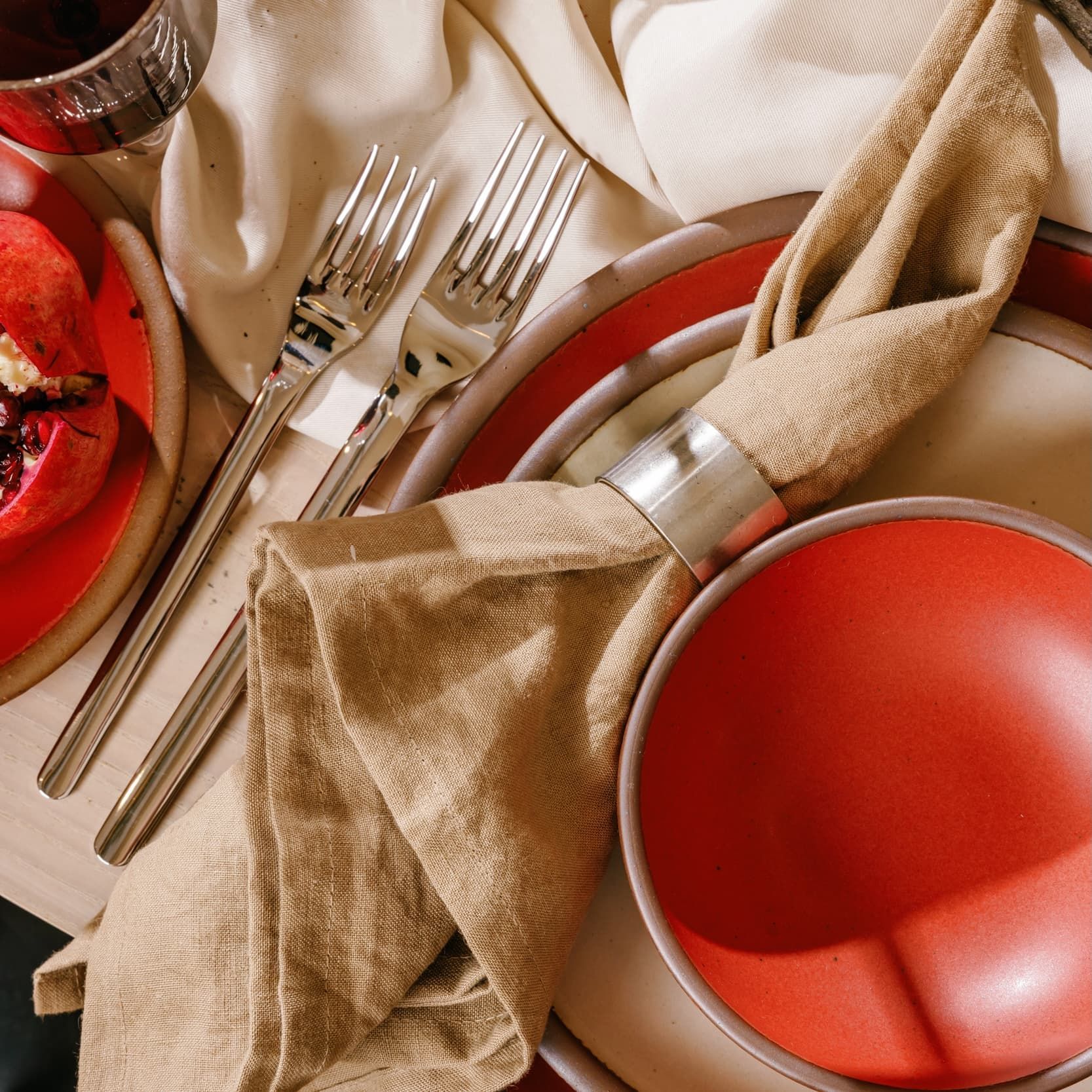 A shiny steel salad fork and dinner fork sit next to a place setting of ceramic dinnerware in cream and bold red colors. There's also a taupe dinner napkin tucked in a pewter napkin ring.
